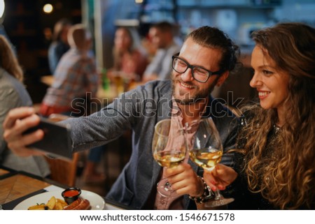 Cute caucasian fashionable couple sitting in restaurant at dinner holding glasses of wine and taking selfie.