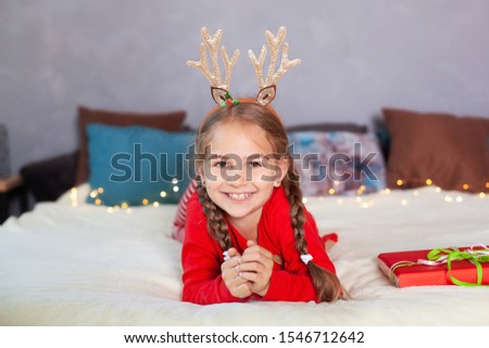Merry Christmas. New Year. Portrait of little smiling girl with christmas gift box at home. cheerful child in red pajamas and with deer horns on his head on Christmas Eve lies on bed and opens gift.