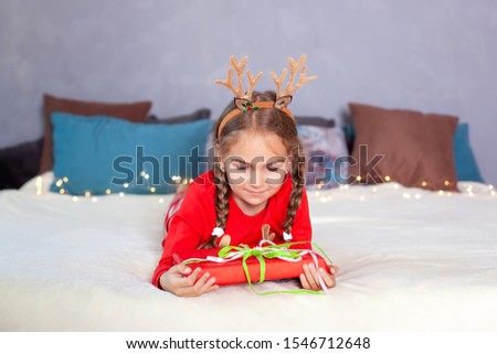 Merry Christmas. New Year. Happy little smiling girl with pigtails with christmas gift box. child in red pajamas and with deer horns on his head lies on bed on Christmas Eve and opens a gift at home.