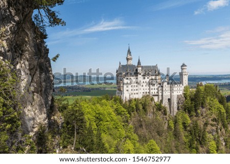 Neuschwanstein castle in Germany from south with a cliff to the left in picture and blue sky and lake forgensee in background. 