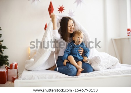 Portrait of happy mother and her baby girl on bed at home with decorated Christmas tree in background