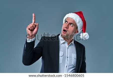 Happy man wearing christmas hat over grey background. Amazed and smiling man while presenting your product and pointing with finger. Christmas promoting, copy space.