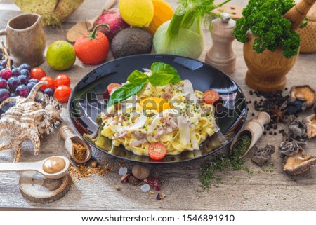 Carbonara pasta with cream, little tomatoes and eggs, presented with vegetables on a wooden table with a black plate.
