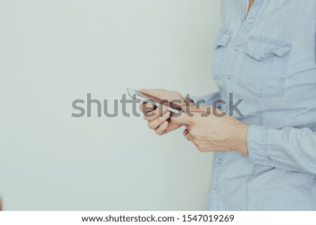 Image of a young woman in a blue shirt working on a modern touchscreen phone at home against a light wall