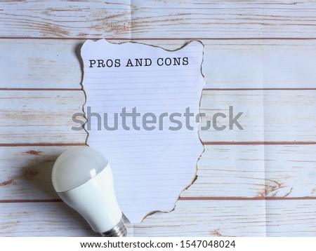 Light bulb and burning word 'PROS AND CONS" in white paper on wooden table. Pros and Cons concept.