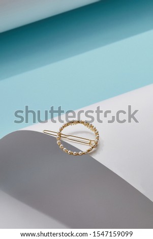 Object photo of a golden round hairpin, decorating with white pearls and metallic beads, lying on a white half-cylinder. The photo was taken on a blue background. 