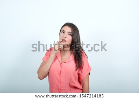 Brunette woman with long hair having doubts while looking up on isolated background. Lady opens her mouth, touches lips with her index finger. Dressed in Pink shirt on white Studio background