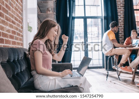 young, happy businesswoman showing winner gesture while sitting on sofa and using laptop near multicultural colleagues