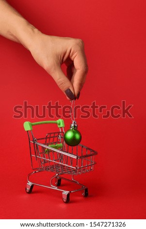 Female hand put green Christmas-tree decorations in a small supermarket trolley on a red background.New Year concept. vertical photo. Place for text.