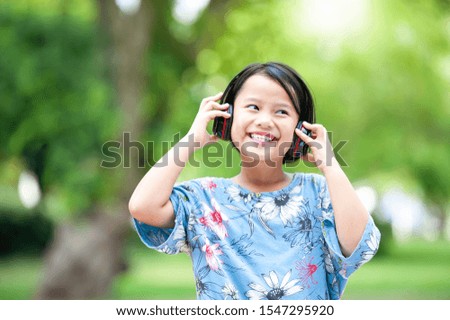 Beautiful Asian young cheerful girl spending good time at the park, listening to music with headphones.Portrait of pretty little girl  listening to music with headphones and smiling in autumn park.