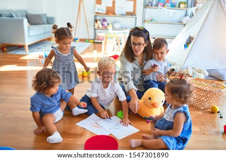 Beautiful teacher and group of toddlers sitting on the floor drawing using paper and pencil around lots of toys at kindergarten Royalty-Free Stock Photo #1547301890