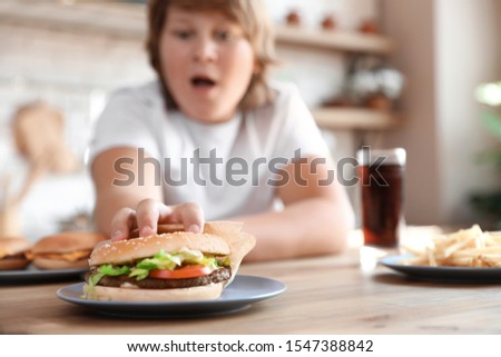 Emotional overweight boy reaching for burger at table in kitchen