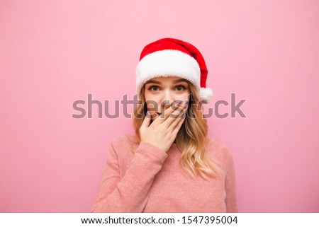 Cheerful girl in a Christmas hat covers her mouth in astonishment, looks into the camera, isolated on a pink background, wears a santa claus hat and a pink sweater. Christmas concept. X-mas