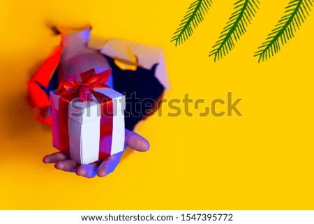 A gift with a red bow in hand emerges from a ragged hole in a yellow paper background, illuminated by neon light