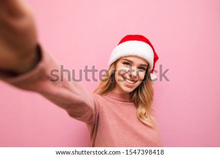 Happy girl in christmas hat makes selfie on pink background, looks into camera and smiles. Portrait of lady in santa hat making her photo on outstretched hand. Christmas selfie concept. X-mas