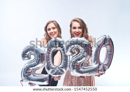 Portrait of happy women in dresses having fun with metallic silver 2020 balloons on white background. New Year celebration and Christmas holidays concept. Copy space.