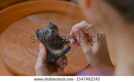 Professional woman potter, decorator painting ceramic souvenir penny whistle toy cat in pottery workshop, studio. Crafting, artwork and handmade concept