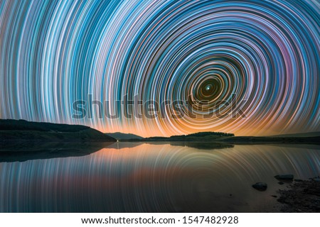 Beautiful night landscape with small lake and mountains. The colorful star trails on the sky. Night time lapse photography.