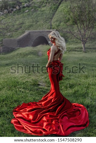 Lady in a tight-fitting red dress with a deep neckline. The princess sat on a rock, her head thrown dreamily. Fantastic shooting. Fashionable tinting. Blonde in a red dress in the forest, satin, gloss Royalty-Free Stock Photo #1547508290