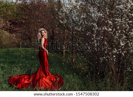 Lady in a tight-fitting red dress with a deep neckline. The princess sat on a rock, her head thrown dreamily. Fantastic shooting. Fashionable tinting. Blonde in a red dress in the forest, satin, gloss Royalty-Free Stock Photo #1547508302