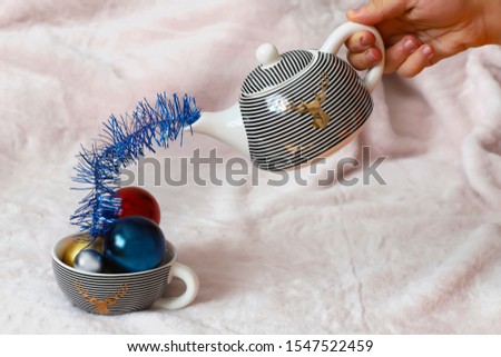 a man's hand from a teapot pours Christmas tinsel into a striped mug inside with Christmas balls on a soft pink background.the unusual concept of the new year
