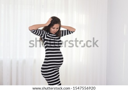 Pregnant woman in striped dress holds hands on belly on a white background. Pregnancy, maternity, preparation and expectation concept. Beautiful tender mood photo of pregnancy.