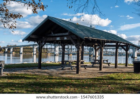 Open shelter in a park with picnic tables along the Kennebec river with a view of a bridge in Bath, Maine on a sunny afternoon with a partly cloudy sky.