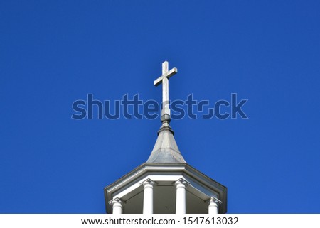 Plain, white Christian cross sits high on top of church spire against blue sky background with copy space.