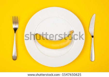 Diet or weight loss concept stock image. One banana on white plate with fork and knife near isolated on yellow background.