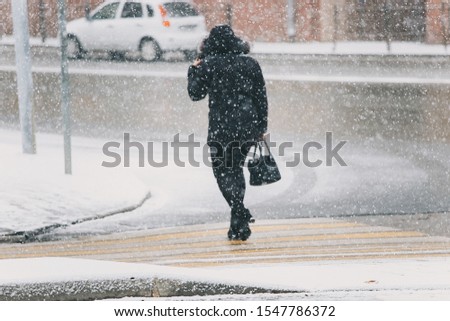 a lone man walks down the street in a heavy snowfall, taking shelter from the flying snow in his face hooded jacket, the first snow in late autumn turned into a storm, cold snap and climate change Royalty-Free Stock Photo #1547786372