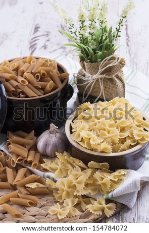 Raw food composition - yellow colored farfelle and brown penne pasta in a clay pot on a bright wooden background.