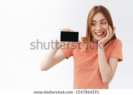 Tender, cute attractive woman in striped t-shirt showing silly, lovely video online, holding smartphone horizontally, gazing mobile phone and sighing from amusement and joy, stand white background