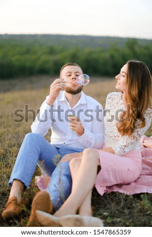 Young guy blowing soap bubbles near caucasian girl and sitting on pink plaid in steppe. Concept of happy couple, romantic love and picnic on nature.