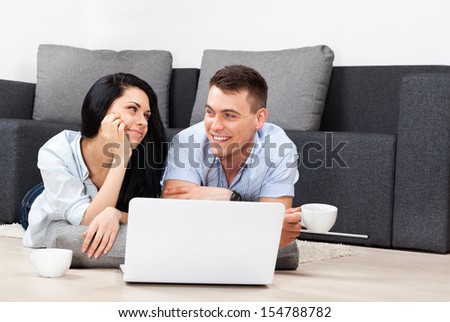 young couple happy smile using laptop, lying on floor near sofa, couch in their living room at home, lovely young man and woman drink coffee hold cup