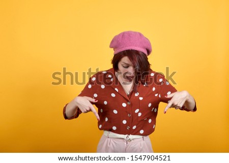 Beautiful caucasian young person wearing funny pink hat over isolated yellow background Pointing down with fingers showing advertisement, surprised face and open mouth. Looking shocked and exciting