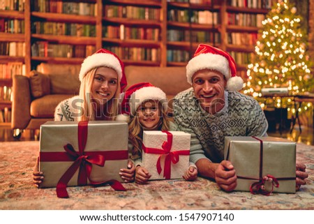 Merry Christmas and Happy New Year! Happy family is waiting for the New Year in Santa Claus hats lie on the floor and hold their gift boxes.