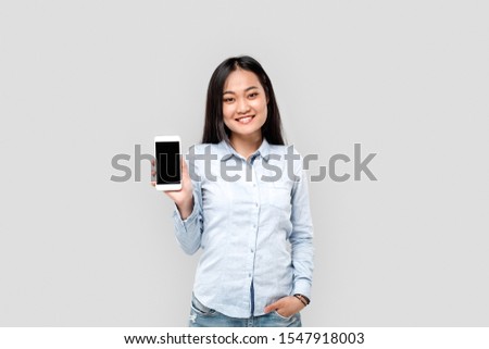 Charming and young adult girl standing isolated on grey background with copy space. Asian female looking at camera and holding in hands smartphone with blank screen