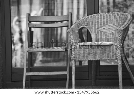  Monochrome photography of wooden chair and wicker outdoor armchair on terrace. Vintage design concept.                        