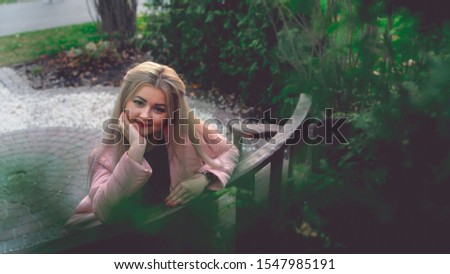 A portrait of a beautiful woman, sitting on a bench near the green vegetation.