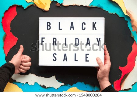 Lightbox text "Black Friday Sale" in burnt paper hole in white, red, yellow and turquoise paper with burned edges, flat lay on black paper with text in light box and hand showing OK sign.