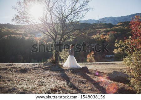 Young beautiful lonely bride posimg on the nature and look to the lake and forest. Female wearing wedding dress and sweater at the background trees against the sunset.