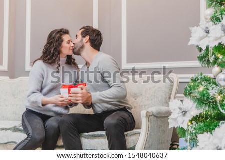 Image of affectionate guy giving present his girlfriend while kissing her, sitting on the sofa in new year time.