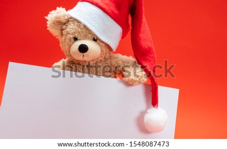 Christmas card template. Teddy bear with santa hat holding a blank paper card, red color background, copy space