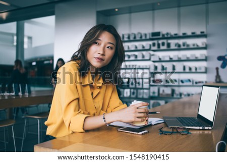 Thoughtful Asian female worker in yellow blouse at desk with laptop on at coffee break in electronics store in Hong Kong city Royalty-Free Stock Photo #1548190415