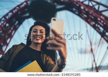 Carefree female vlog maker smiling at front camera while shooting selfie video for sharing to social networks via smartphone gadget, happy woman in optical spectacles clicking photo pictures