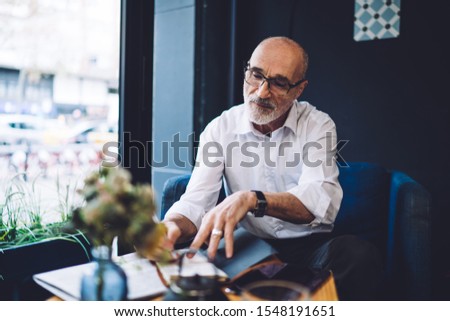 Elderly bald 60 years old gray bearded businessman with eyeglasses in white shirt sitting at table on blue chair and signing documents in cafe 