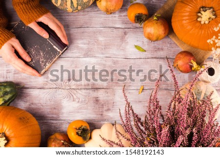 Autumn thanksgiving moody background with different pumpkins, fall fruit and flowers on rustic wooden table. Flat lay with female hands holding a book