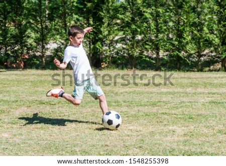 Child playing football in a stadium. Trees on the background. Casual clothes. Kid play with ball. Sunny day on green meadow. Little boy playing football. Concept for sport and recreation.