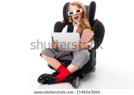 A little girl with makeup and long blonde hair sitting in a car baby chair with tablet, earphones, pop-corn, 3D glasses and watch interesting film of cartoon