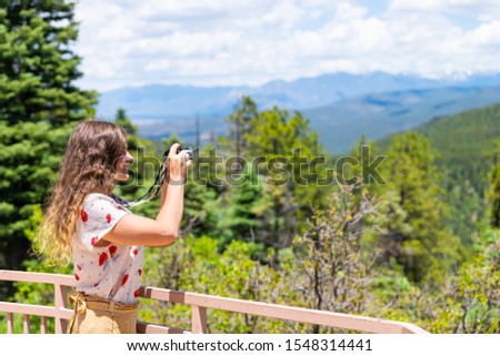 Woman taking picture of Carson National Forest green pine trees with Sangre de Cristo mountains on summer peak overlook from route 76 high road to Taos